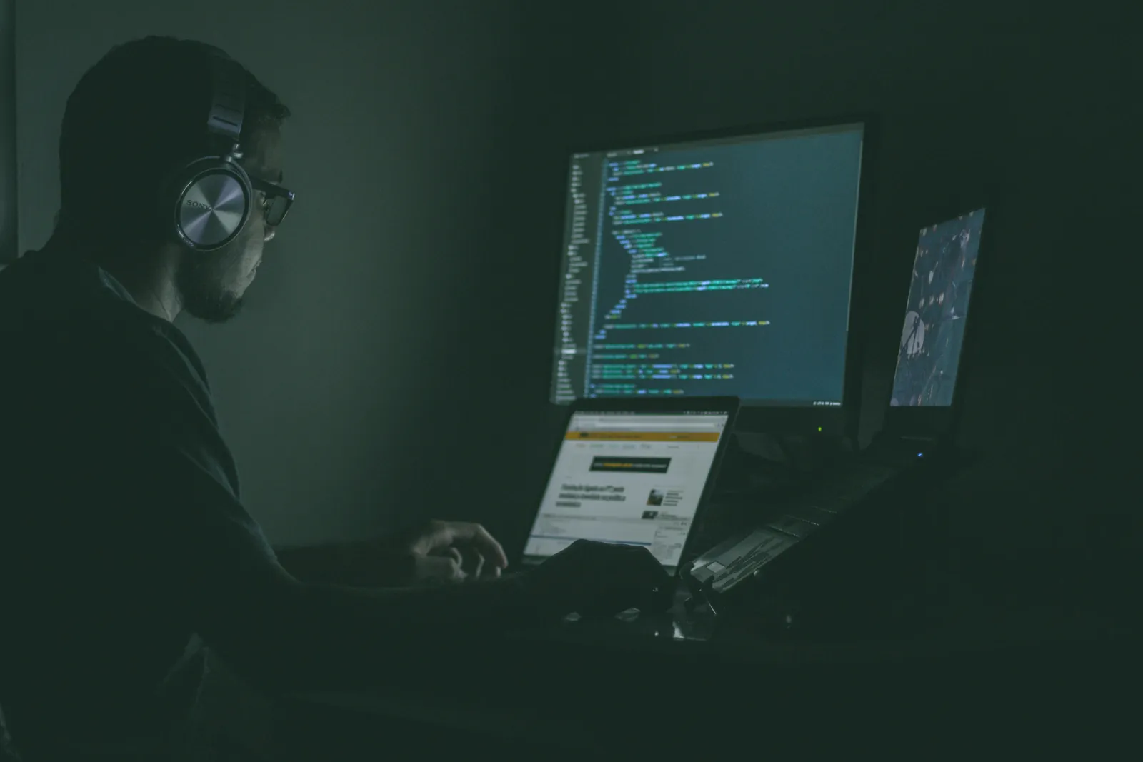 A developer in headphones working at a laptop in a dark room, code on the monitor behind